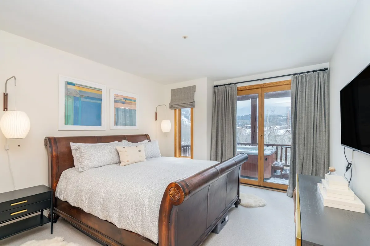 This image features a sleigh bed set against large windows that flood the room with natural light. There's subtle artwork on the wall and a television, creating a cozy and serene bedroom retreat ideal for relaxation and privacy.