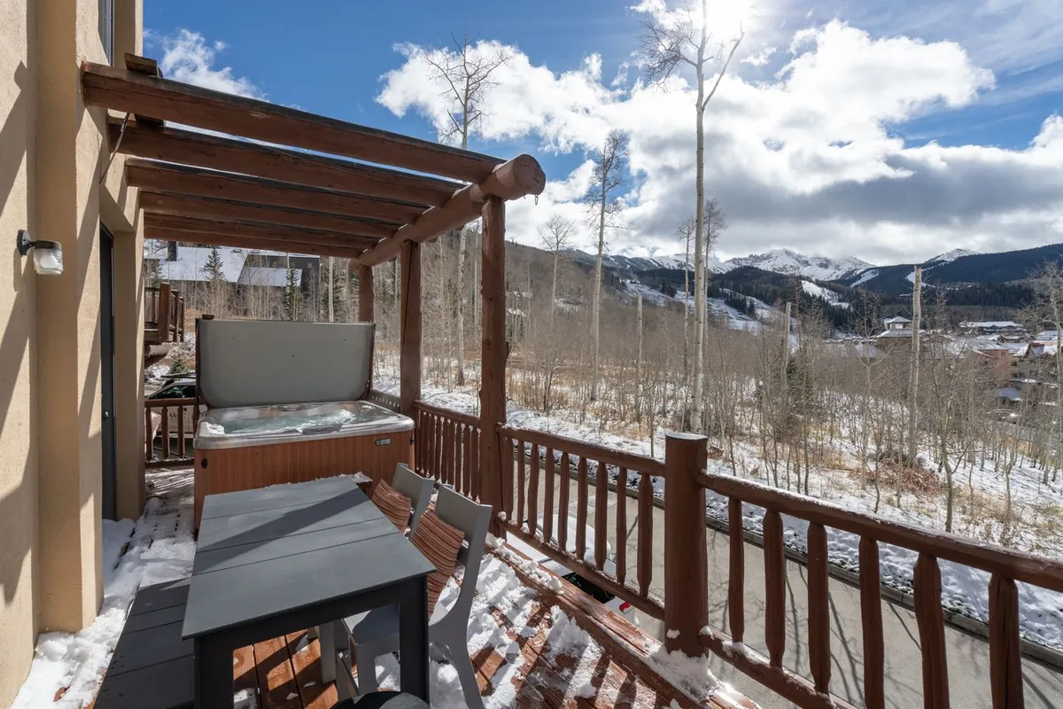 An expansive deck offering both a hot tub and an outdoor dining table, all set against a panoramic mountain backdrop. Wooden railings frame the view, inviting guests to enjoy fresh alpine air and the seamless indoor-outdoor transition.