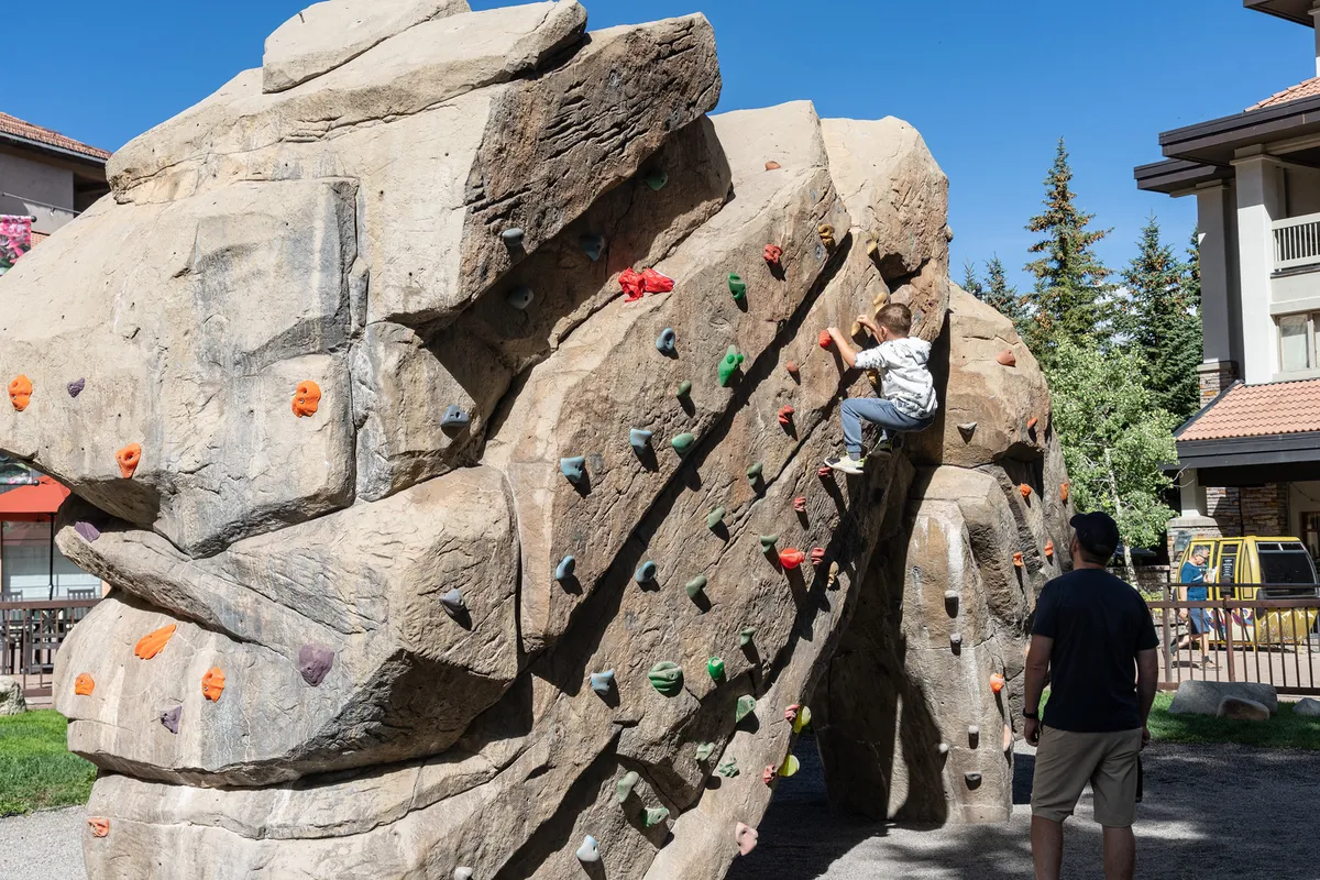 A child climbs a rock wall outdoors under the watchful eye of an adult, set against a backdrop of trees and clear skies. The scene highlights family-friendly adventure and safe outdoor play opportunities.