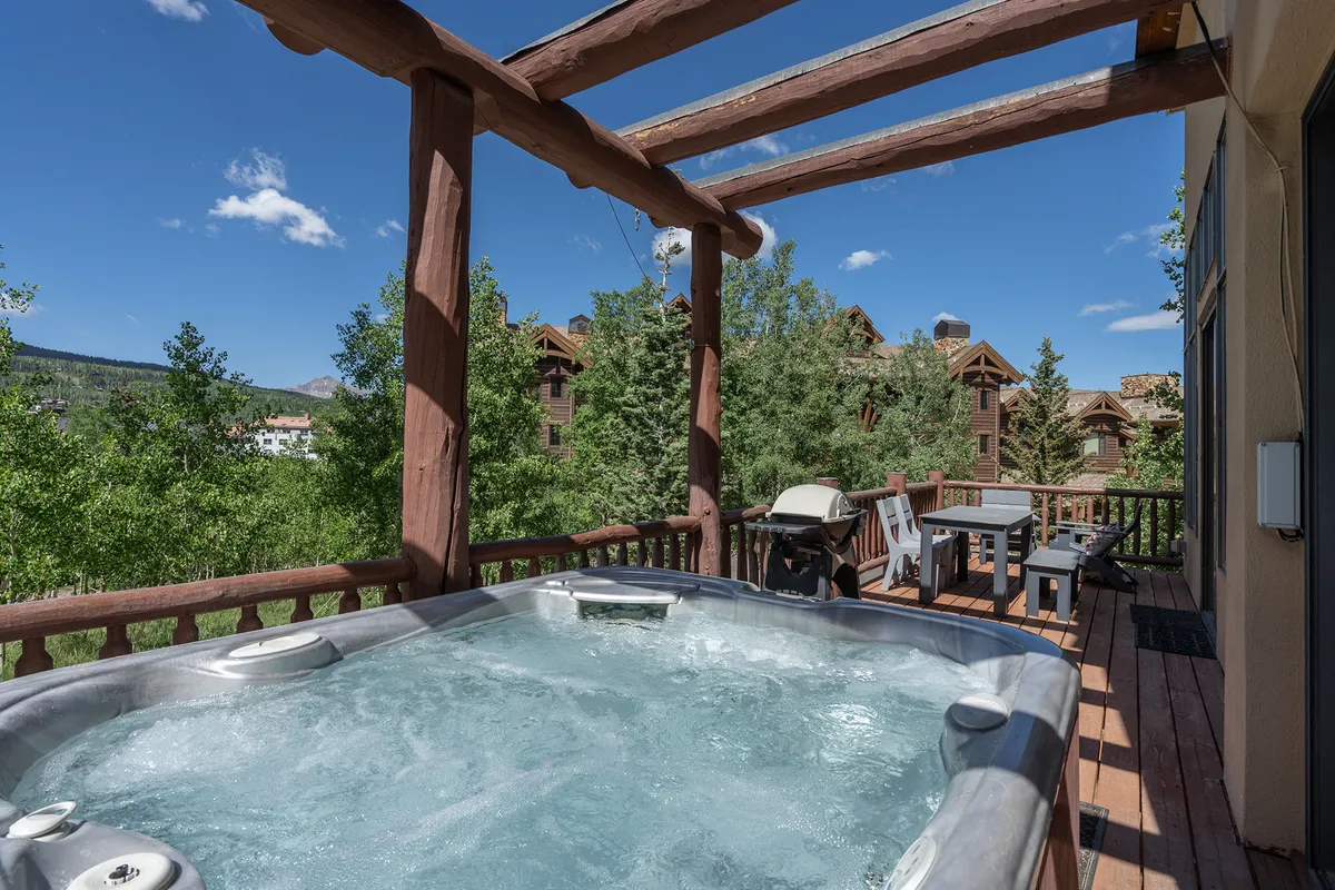 A generously sized hot tub sits on a wooden deck surrounded by majestic trees, with comfortable outdoor dining space visible nearby. The ambiance is tranquil yet inviting, hinting at both relaxation and outdoor social gatherings after a day of exploring.