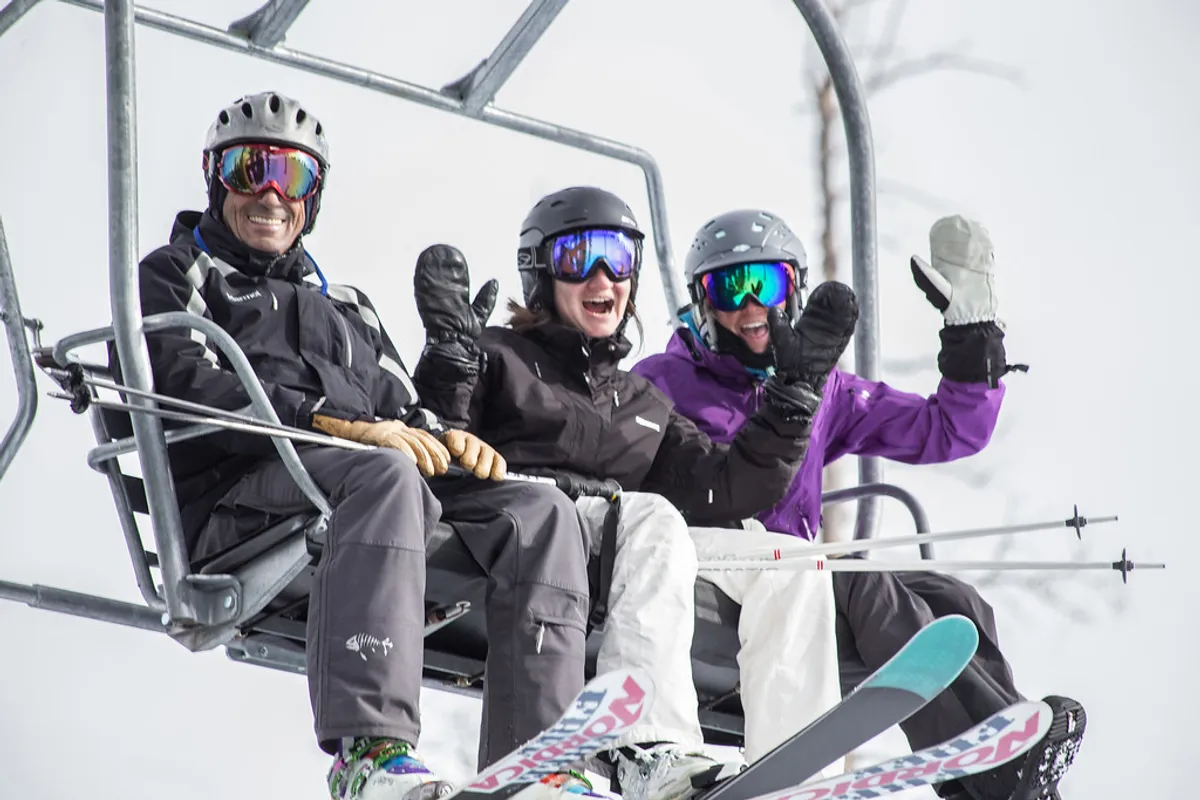 Three skiers are riding a chairlift above a snow-covered mountain landscape, ski equipment in hand and smiles on their faces, set against a bright, crisp winter scene. Their camaraderie and excitement are evident, highlighting the social and adventurous spirit of a mountain getaway.