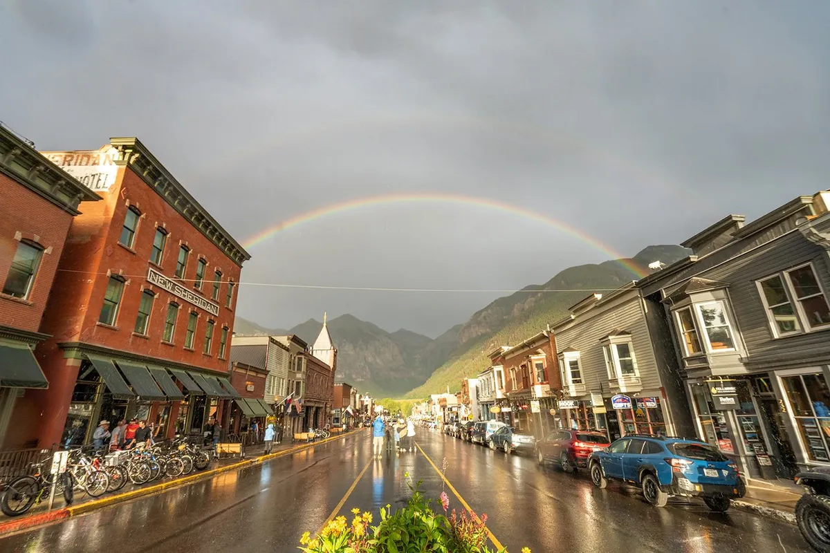 A lively yet intimate view of Mountain Village’s main street, lined with historic buildings and boutique shops. A rainbow arches above bicyclists and strolling pedestrians, adding a whimsical touch to the upscale, vibrant village atmosphere.