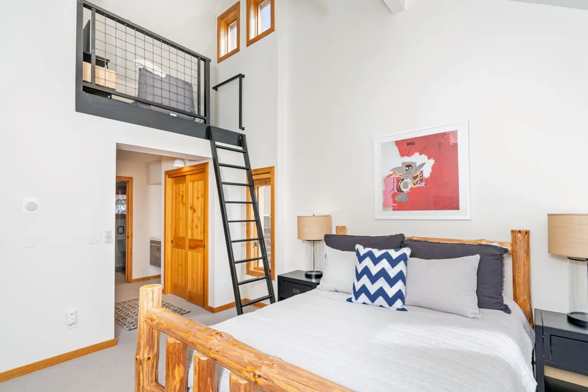 A comfortable bedroom featuring a wooden bed frame, a ladder leading to a lofted sleeping space above, and welcoming decor. Large windows provide natural light, and the artwork adds a touch of charm.