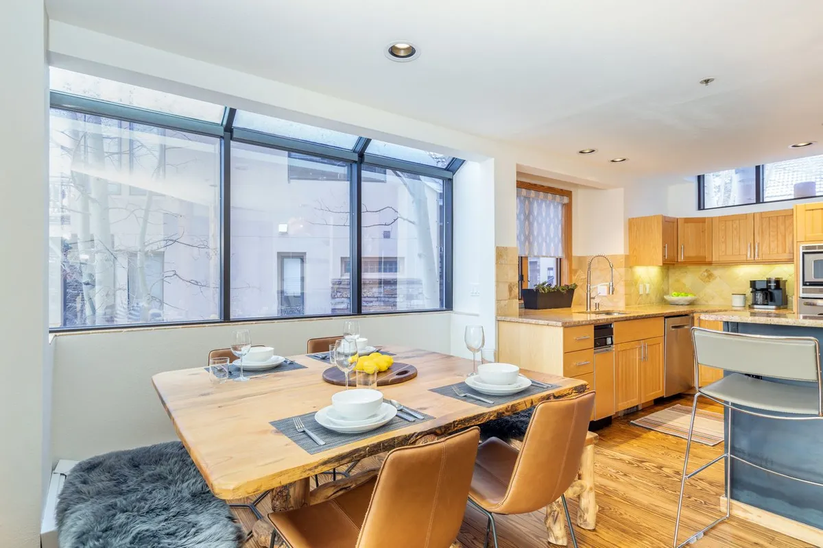 A dining area with a spacious table set for a meal, flanked by large windows that provide sweeping views of the surrounding mountains. The adjacent kitchen counter hints at seamless flow for preparing and serving meals. Abundant natural light fills the space, creating a welcoming atmosphere for gatherings.