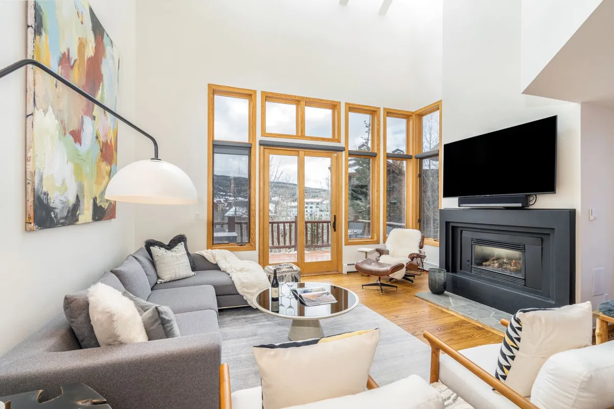 The hero image showcases a stunning, contemporary living room featuring expansive windows that frame mountain vistas, chic modern furnishings, a glowing fireplace, and bold abstract artwork. The inviting space is anchored by a round coffee table, creating the perfect setting for relaxation and socializing.