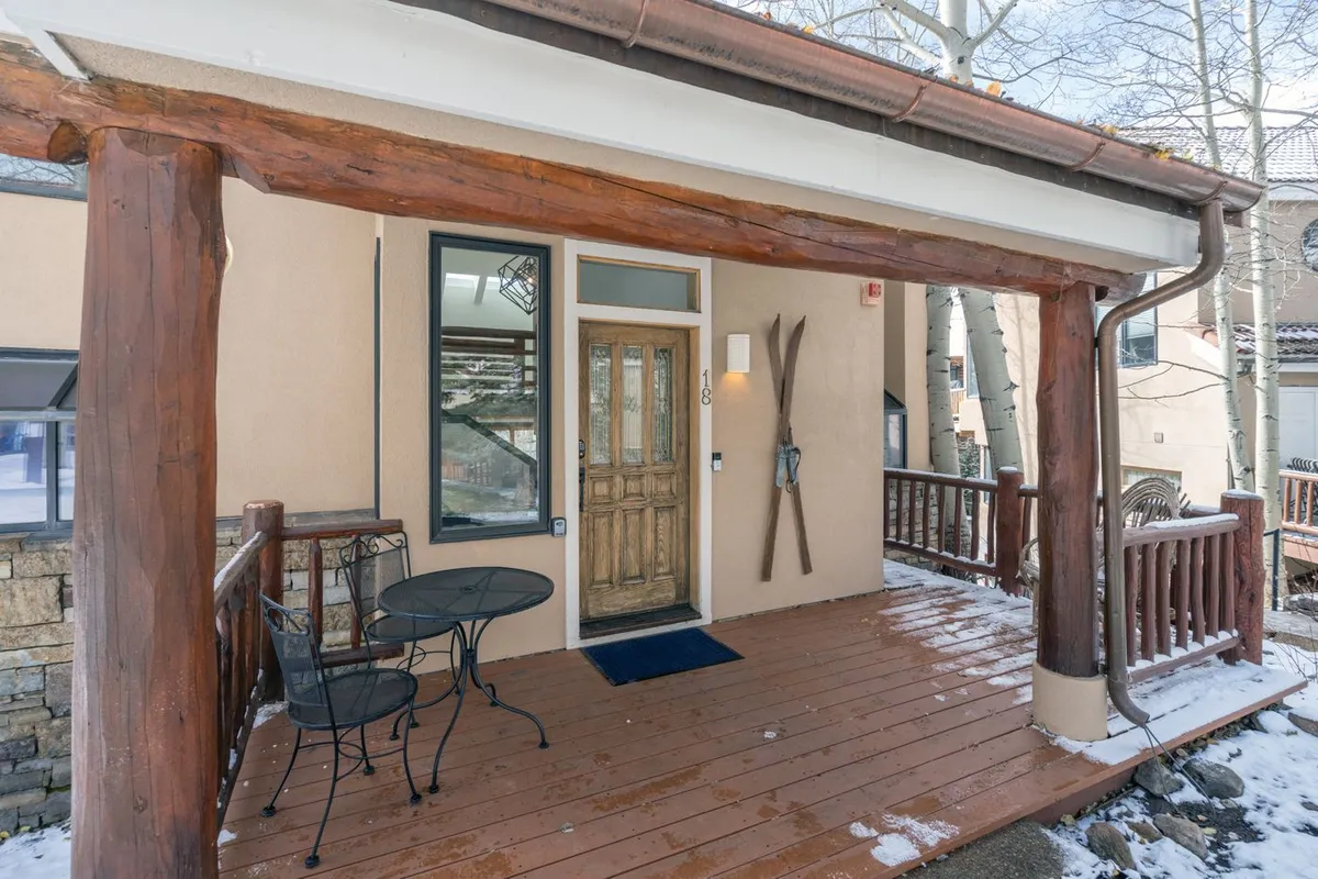 The shot displays a wooden porch with a front door, small table and chairs, and ski equipment decor. The setting suggests a cozy, inviting atmosphere for relaxing after a day of outdoor activities and emphasizes accessibility for adventure gear.